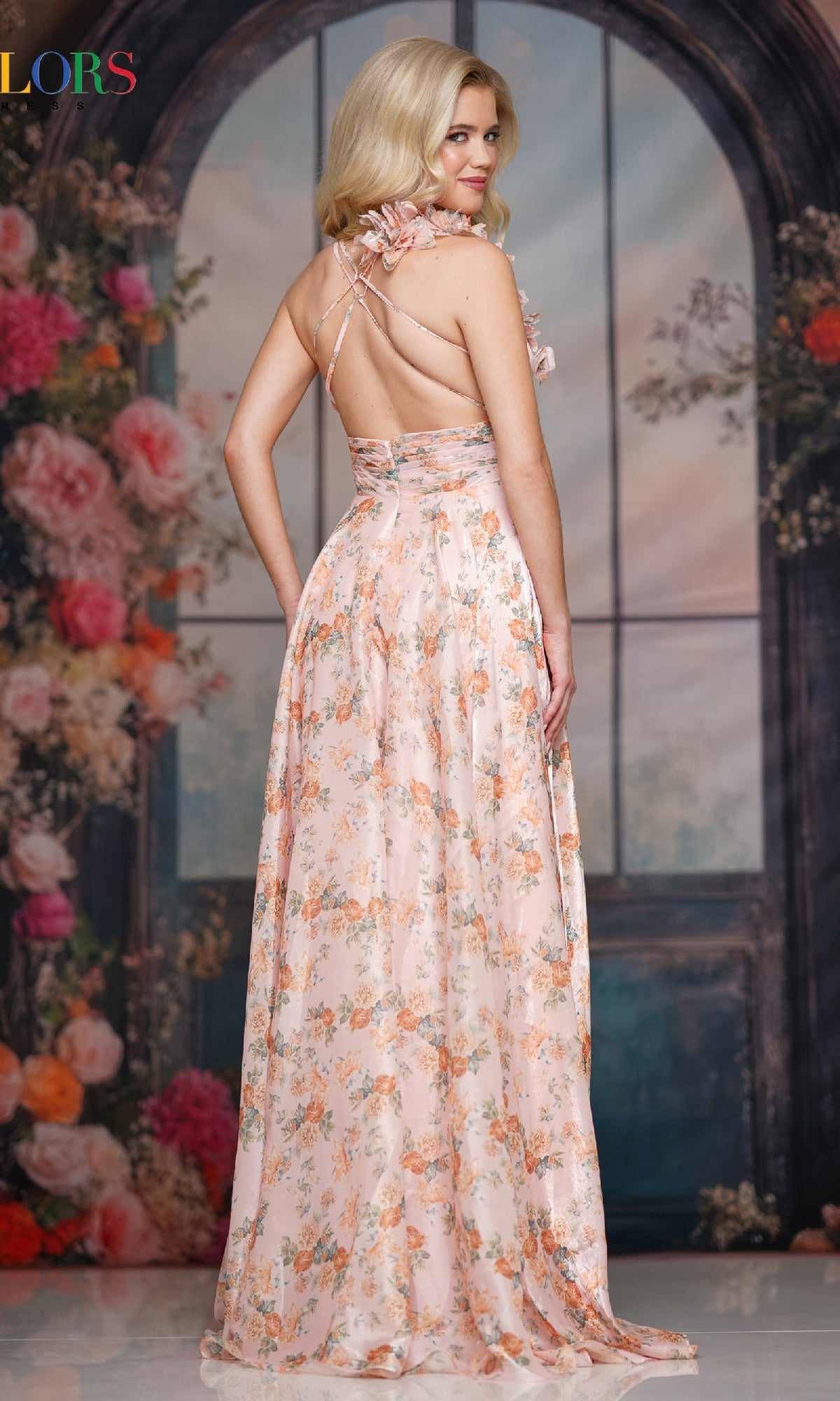 A woman models the Colors Dress 3807, a long satin print prom dress, posing with her back turned before a decorative backdrop with flowers and arched windows.